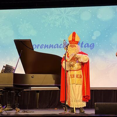 Bild vergr&ouml;&szlig;ern: Drei Personen auf einer B�hne: eine wei� gekleidete Pianistin am Fl�gel, ein Nikolaus und ein Mann mit einem Mikrofon