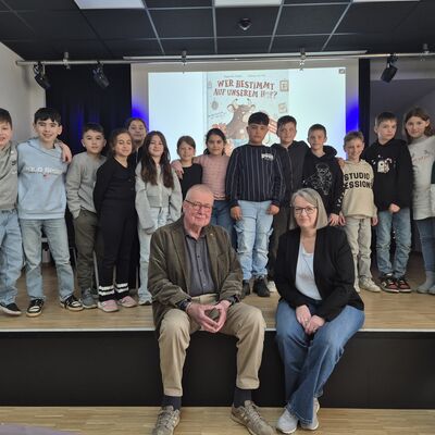 Bild vergr&ouml;&szlig;ern: Zwei Erwachsene sitzend auf einer B�hne, dahinter einige Kinder.