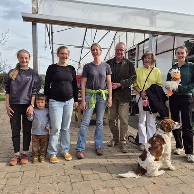 Bild vergr&ouml;&szlig;ern: Acht Erwachsene, ein Kind und ein Hund auf einem Gruppenfoto vor einem Gew�chshaus.